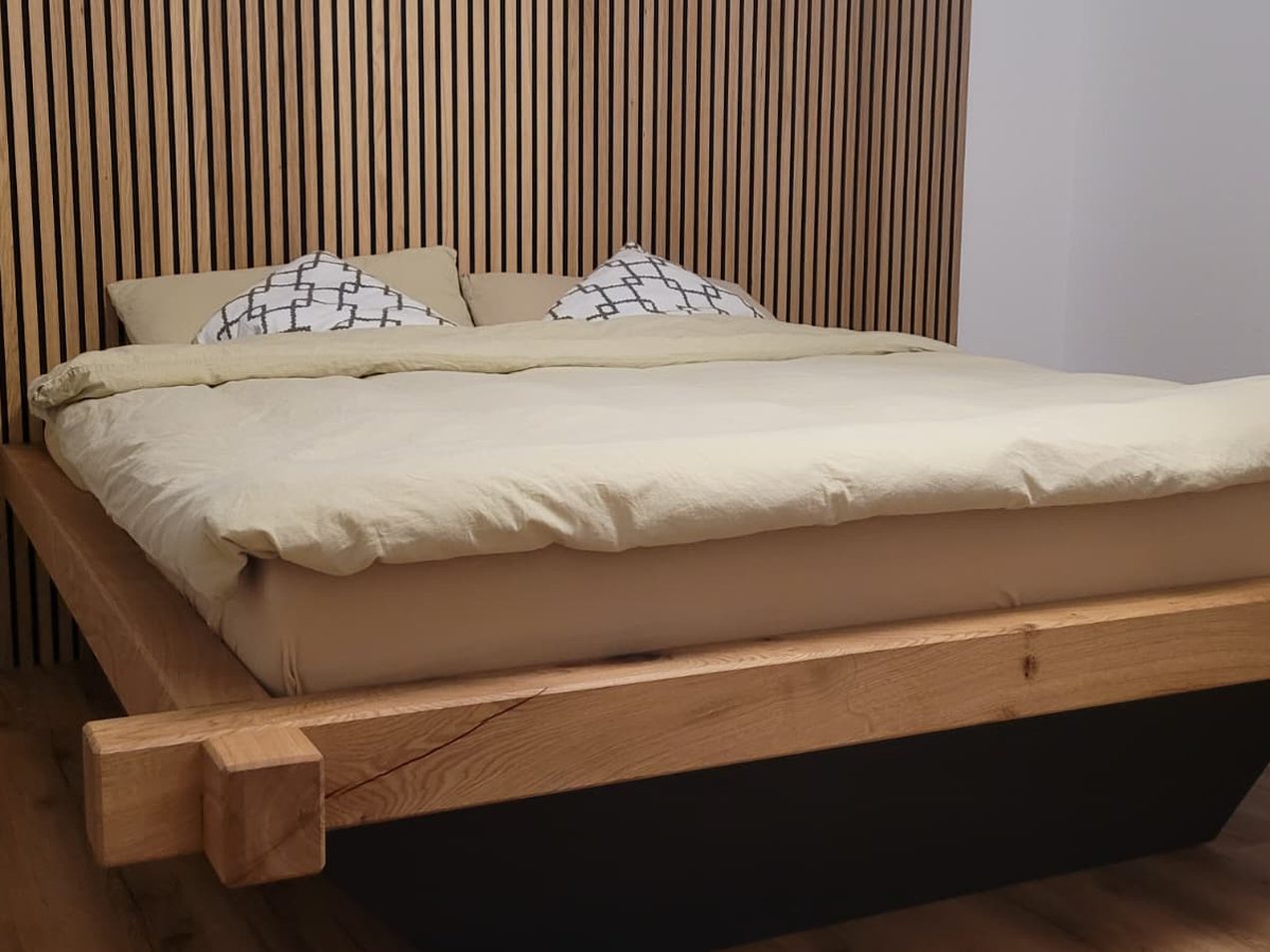 Massivholzbett aus kräftigen Holzbalken vor einer Wand mit Holzlamellen